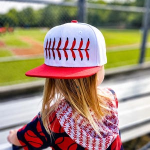 May include: White and red baseball cap with a red brim. The cap features a baseball stitch design in red. The person in the image is wearing a baseball-themed hoodie with red baseballs on a navy blue background.