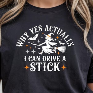 Può includere: T-shirt nera con la scritta "WHY YES ACTUALLY I CAN DRIVE A STICK" e una grafica di una strega su una scopa. Il design include pipistrelli e stelle, perfetto per Halloween.