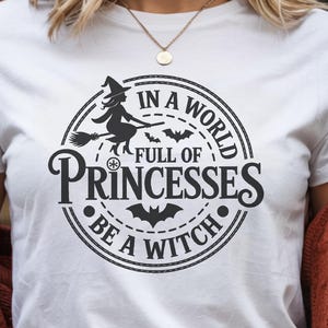 Puede incluir: Camiseta blanca con un gráfico circular negro que dice "In a world full of princesses be a witch". El diseño incluye una bruja en una escoba y murciélagos. La persona lleva un suéter color óxido.