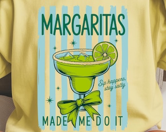 May include: A light yellow sweatshirt with the words "MARGARITAS" in green, and a graphic of a margarita cocktail. The design includes a lime slice, ice, and a green bow. The text "Sip happens, stay salty" and "MADE ME DO IT" are also included.