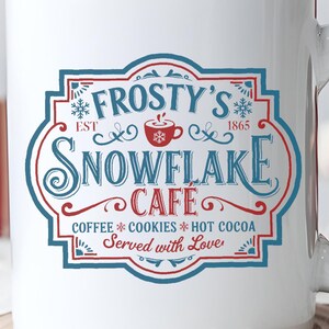 Puede incluir: Taza de cerámica blanca con el texto "Frosty's Snowflake Cafe" en un diseño azul y rojo. La taza también presenta las palabras "Coffee, Cookies, Hot Cocoa, Served with Love". La taza está sobre una superficie de madera.