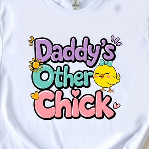 Puede incluir: Camiseta blanca con el texto "Daddy's Other Chick" en letras de burbujas coloridas, con un pollito de dibujos animados con un lazo azul. El diseño incluye un sol y corazones. La camiseta se muestra sobre una superficie blanca.
