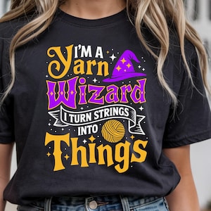 May include: Black t-shirt with the text "I'M A Yarn Wizard I Turn Strings Into Things" in purple and gold. The design includes a witch hat and a ball of yarn. The shirt is paired with blue denim shorts.