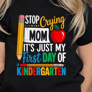 Puede incluir: Una camiseta negra con el texto "STOP Crying MOM IT'S JUST MY First DAY OF KINDERGARTEN" en letras coloridas. El diseño incluye un lápiz amarillo, una manzana roja y un lápiz. La camiseta la lleva un niño pequeño.
