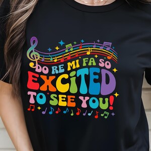以下が含まれることがあります： カラフルな音楽デザインの黒いTシャツ。「Do Re Mi Fa So Excited To See You!」のテキストが虹色で書かれています。音符とト音記号もデザインにあります。このシャツは音楽をテーマにしたアパレルです。