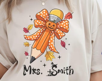 May include: Beige t-shirt with a pencil design, pumpkin bow, and autumn leaves. The text "Mrs. Smith" is printed below. The shirt is worn with light blue denim shorts.