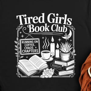 Op de afbeelding: Zwart T-shirt met een witte afbeelding met de tekst "Tired Girls Book Club". Het ontwerp bevat een open boek, een kopje koffie, kaarsen en een kleine plant. De tekst "Running on Coffee, Chaos + Chapters" is ook inbegrepen.