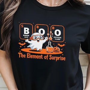 May include: Black t-shirt featuring a Halloween-themed graphic. The design includes the word "BOO" in periodic table elements, a cartoon ghost, a pumpkin, and the text "The Element of Surprise".