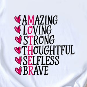 May include: White t-shirt with the word "MOTHER" spelled out vertically in black letters, each letter preceded by a pink heart. The words "AMAZING", "LOVING", "STRONG", "THOUGHTFUL", "SELFLESS", and "BRAVE" are also displayed.