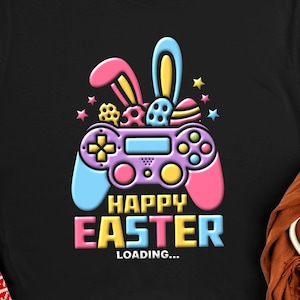 Puede incluir: Camiseta negra con un gráfico colorido que presenta un mando de videojuegos con orejas de conejo y huevos de Pascua. El texto "HAPPY EASTER LOADING..." se muestra en colores vibrantes. Un diseño festivo para Pascua.
