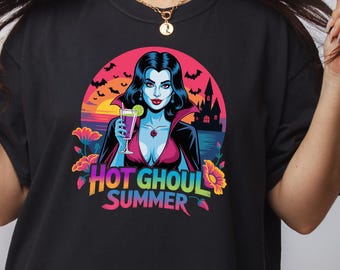 May include: Black t-shirt featuring a colorful graphic of a vampire woman holding a cocktail, with the text "HOT GHOUL SUMMER". The design includes a sunset, bats, and a castle. The shirt is a comfortable, casual style.