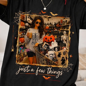 May include: A black t-shirt featuring a rectangular graphic print of a person in a skeleton shirt shopping in a store filled with Halloween decorations, including pumpkins and a skeleton. The text "just a few things" is printed below the graphic.