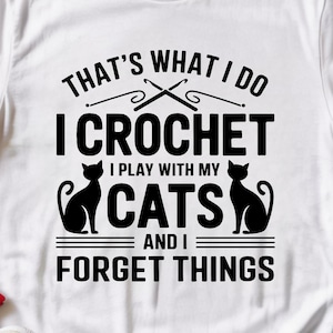 May include: White t-shirt featuring the text "THAT'S WHAT I DO I CROCHET I PLAY WITH MY CATS AND I FORGET THINGS" in black. The design includes crochet hooks and two black cat silhouettes.