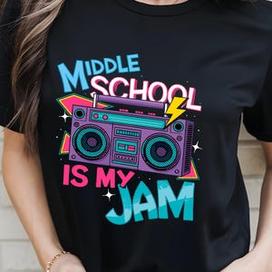 May include: Black t-shirt with a colorful graphic design. The design features a retro boombox with the text "MIDDLE SCHOOL IS MY JAM" in teal, pink, and purple. A yellow lightning bolt is also included in the design.