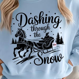 May include: Light blue sweatshirt with a black graphic of a horse-drawn sleigh and the text "Dashing through the Snow." The design includes trees and dots. The sweatshirt is a crew neck style.