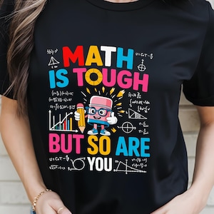 May include: Black t-shirt with the text "MATH IS TOUGH BUT SO ARE YOU" in colorful letters. The design includes a cartoon eraser character holding a pencil, surrounded by math symbols and equations. The shirt is a casual wear item.