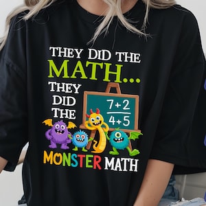 Puede incluir: Camiseta negra con el texto "THEY DID THE MATH..." en blanco y verde. La camiseta presenta una caricatura de monstruos y una pizarra con problemas de matemáticas. La frase "MONSTER MATH" está en amarillo.