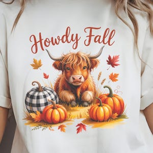 May include: Cream-colored t-shirt featuring a Highland cow illustration with the text "Howdy Fall." The design includes pumpkins, fall leaves, and a buffalo plaid pumpkin. The image has a fall theme.