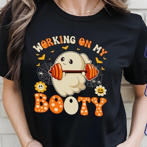 May include: Black t-shirt with a Halloween-themed graphic. The design features a cartoon ghost lifting weights with the text "WORKING ON MY BOOTY" in orange and white. Includes bats, spiderwebs, and flowers.