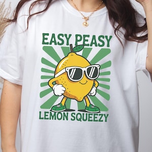 May include: White t-shirt featuring a cartoon lemon wearing sunglasses, with the text "EASY PEASY LEMON SQUEEZY" in green. The lemon is yellow with green shoes and gloves. The design has a green sunburst background.