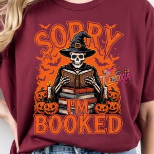 Peut inclure: Un t-shirt bordeaux avec un motif de squelette portant un chapeau de sorcière, lisant un livre devant une pile de livres, des bougies et des citrouilles d'Halloween. Le texte SORRY I'M BOOKED est écrit en orange sur un fond rempli de chauves-souris.
