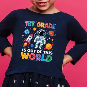 May include: Navy blue long-sleeve shirt with a colorful graphic featuring an astronaut, planets, and a rocket. The text reads "1st Grade is Out of This World". The shirt is part of a back-to-school collection.