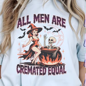 Puede incluir: Camiseta azul claro con un diseño de Halloween. El diseño presenta una bruja, un esqueleto en un caldero, murciélagos y el texto "ALL MEN ARE CREMATED EQUAL". El diseño utiliza tonos rojos, naranjas y negros.