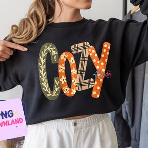 Peut inclure: Un sweat-shirt noir à manches longues avec le mot COZY imprimé en grandes lettres stylisées sur la poitrine. Chaque lettre présente un motif unique : le C est vert olive avec des feuilles, le O et le Z sont orange avec des fleurs blanches, et le Y est orange avec des pois blancs.