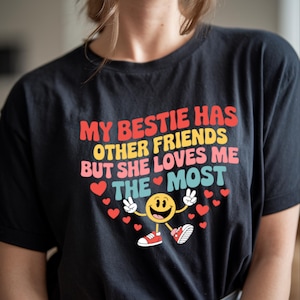 May include: A black t-shirt with a colorful graphic that says "My bestie has other friends but she loves me the most". The graphic includes a smiling face with peace signs and red hearts.