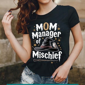 May include: Black t-shirt featuring a graphic of a witch's hat, an owl, and a wand. The text "MOM Manager of Mischief" is printed on the shirt.