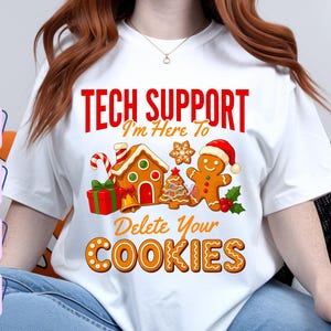 Puede incluir: Una camiseta blanca con un gráfico que incluye el texto rojo TECH SUPPORT, el texto naranja I'm Here To y el texto naranja Delete Your COOKIES. El diseño incluye una casa de jengibre, un muñeco de jengibre y motivos navideños.