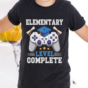 Puede incluir: Camiseta negra con un diseño gráfico que presenta un mando de videojuegos, un birrete y la frase "ELEMENTARY LEVEL COMPLETE". El diseño incluye pergaminos y estrellas, ideal para una celebración de graduación.