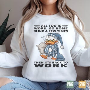 Op de afbeelding: Witte sweatshirt met een cartoon eend met een slaapmuts en pyjama, met een kussen. Tekst luidt: "ALL I DO IS WORK, GO HOME BLINK A FEW TIMES THEN IT'S BACK TO WORK."