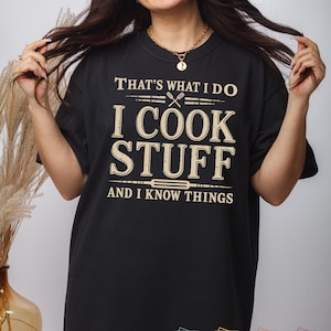 May include: Black t-shirt with the text "THAT'S WHAT I DO, I COOK STUFF, AND I KNOW THINGS" in a tan color. The design includes crossed utensils and a rolling pin graphic. The shirt has short sleeves.