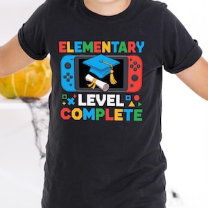 May include: Black t-shirt with a colourful graphic that reads "ELEMENTARY LEVEL COMPLETE." The design features a blue and red game controller with a graduation cap and diploma. The shirt is worn by a person with face paint.