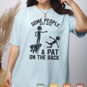 Może przedstawiać: Jasnoniebieska koszulka z czarnym napisem "SOME PEOPLE JUST NEED A PAT ON THE BACK". Grafika przedstawia ludzika na klifie wyciągającego rękę do drugiego spadającego. Koszulka ma krótkie rękawy.