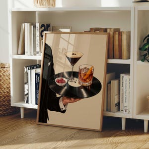 Pode incluir: Uma impress&atilde;o art&iacute;stica emoldurada que mostra uma pessoa com um casaco de fato preto a segurar um disco de vinil preto como tabuleiro. No disco est&atilde;o um copo de martini com gr&atilde;os de caf&eacute;, um copo de u&iacute;sque com casca de laranja e uma pequena ta&ccedil;a com cerejas vermelhas.