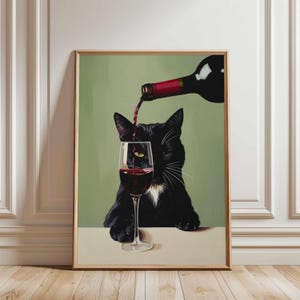 Op de afbeelding: Een ingelijste kunstprint toont een zwarte kat met witte accenten, die kijkt naar rode wijn die in een glas wordt geschonken. De achtergrond is groen en de lijst lichtbruin. Humoristisch thema.