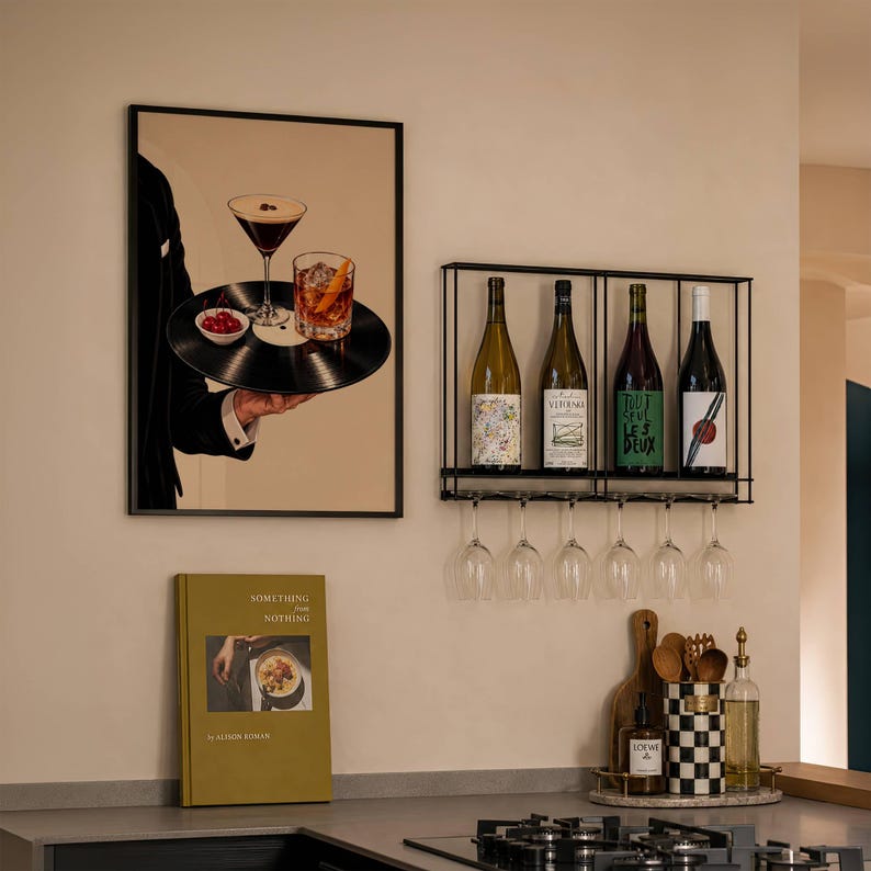 Pode incluir: Uma impress&atilde;o de parede emoldurada mostra uma pessoa de fato preto segurando uma bandeja de disco de vinil com um martini, um cocktail e cerejas. Ao lado, um suporte de metal preto cont&eacute;m quatro garrafas de vinho sobre seis ta&ccedil;as.