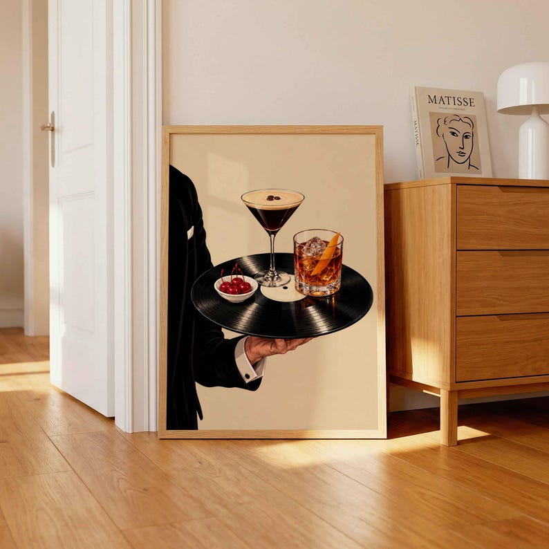 Pode incluir: Uma impress&atilde;o de arte emoldurada mostrando uma pessoa com um palet&oacute; preto segurando um disco de vinil preto como bandeja. No disco, h&aacute; uma ta&ccedil;a de martini com gr&atilde;os de caf&eacute;, um copo de u&iacute;sque com gelo e casca de laranja e uma pequena tigela.