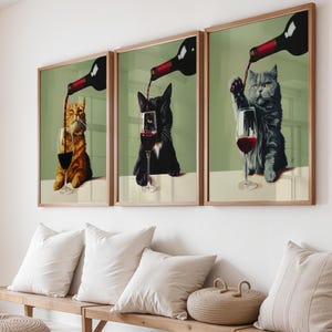 以下が含まれることがあります： ワインをグラスに注ぐ猫が描かれた、額入りのアートプリント3枚。猫はオレンジ、黒、グレーで、それぞれ赤ワインのグラスを持っています。背景は淡い緑色で、フレームは木製です。