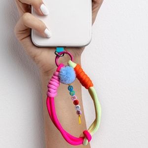 Può includere: Uno smartphone bianco con un cinturino da polso colorato. Il cinturino presenta segmenti di corda rosa neon, arancione e verde chiaro, un piccolo pon-pon azzurro e un ciondolo con perline che compongono il nome CAROEN in lettere colorate.