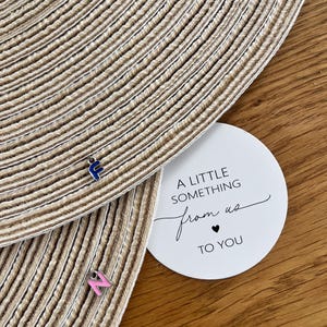 Può includere: Primo piano di un sottobicchiere beige intrecciato con un ciondolo a forma di lettera "E" blu e un ciondolo a forma di lettera "N" rosa. Un'etichetta rotonda bianca recita "A LITTLE SOMETHING from us TO YOU" in corsivo nero.