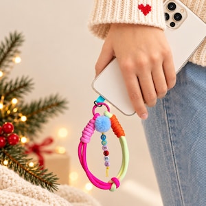 Könnte beinhalten: Ein Handy-Armband mit einer leuchtenden Schlaufe aus neonpinker, oranger und hellgrüner Kordel. Es enthält einen kleinen hellblauen Pompon und einen Anhänger mit bunten Buchstabenperlen, die den Namen CAREN bilden, ergänzt durch eine rote Perle.