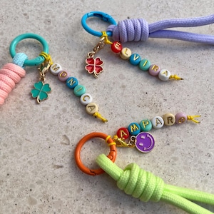 Può includere: Tre portachiavi con cordini intrecciati colorati in rosa, lavanda e verde lime attaccati ad anelli di metallo. Ogni portachiavi include un ciondolo, come un quadrifoglio o una faccina sorridente, e perline che compongono i nomi Monica, Elvira e Amparo.