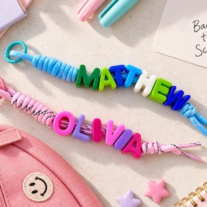 Pode incluir: Dois puxadores de zíper ou chaveiros personalizados feitos de paracord e contas de letras de plástico. Um apresenta o nome MATTHEW em contas verdes, azuis e brancas em um cordão azul. O outro apresenta o nome OLIVIA em contas rosas e roxas em um cordão.