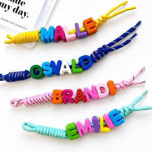 Può includere: Quattro braccialetti con nomi colorati con i nomi MALLE, OSVALD, BRANDI ed EMILIE scritti in lettere maiuscole luminose. Ogni braccialetto presenta un cordino di colore diverso e colori delle lettere. È visibile anche una piccola carta con il testo "Your order made my day. THANK YOU SO MUCH!"