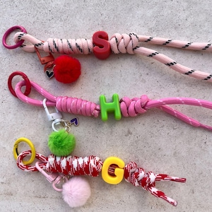 Può includere: Tre portachiavi fatti a mano con corde colorate e perline con lettere. Un portachiavi è rosa con una perlina "S" rossa, un altro è rosa con una perlina "H" verde, e l'ultimo è rosso e bianco con una perlina "G" gialla. Ognuno ha un pompon.