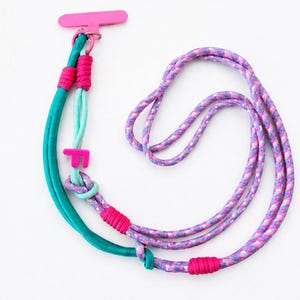 Op de afbeelding: Een kleurrijk telefoonkoord met een roze telefoonhouder. Het koord heeft teal, lichtblauwe en roze koorden. Het ontwerp bevat geknoopte secties en een roze letter "F" accent, wat een levendig accessoire creëert.