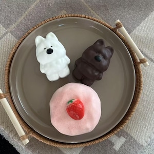 May include: A decorative plate with a white bear-shaped figurine, a brown bear-shaped figurine, and a pink pastry topped with a strawberry. The plate is set on a woven tray.